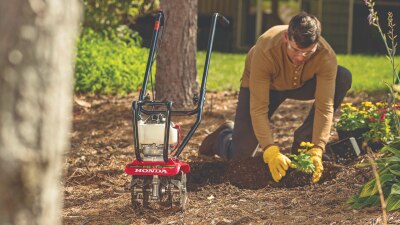 A gardener, on their knees, plants flowers behind a Honda tiller. 