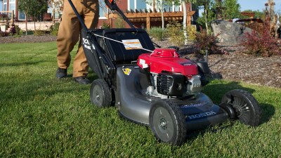 A landscaper pushes a Honda mower near a bush on a golf course. In the background is another landscaper and a golfer. 