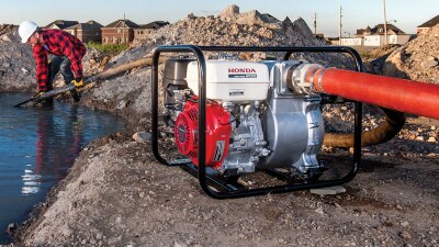 Closeup of a Honda construction pump on the edge of water. In the background, a construction worker holds a hose in the water.  