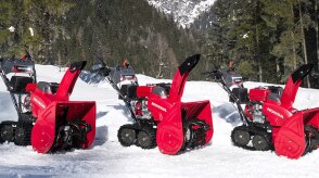 Wide view of three snowblowers parked side by side on a bed of snow with a coniferous forest in the background.  