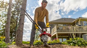 Wide view of homeowner with gloves and protective eyewear lifting a tiller in his yards; house in background.  